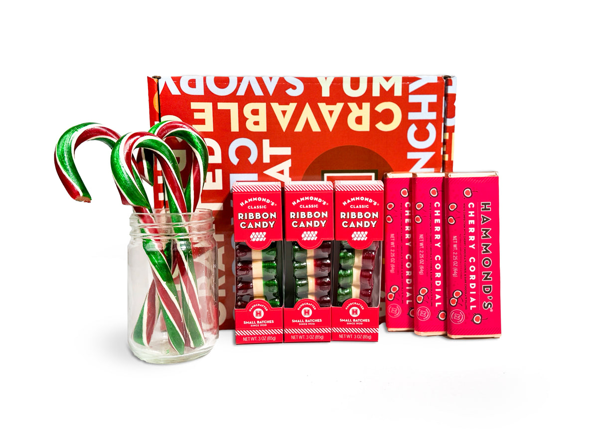 A holiday candy gift set featuring a clear glass jar filled with red and green striped candy canes, several small red boxes of assorted candies, and a tall red box of cherry cordials, arranged in front of a large red branded package on a white background.