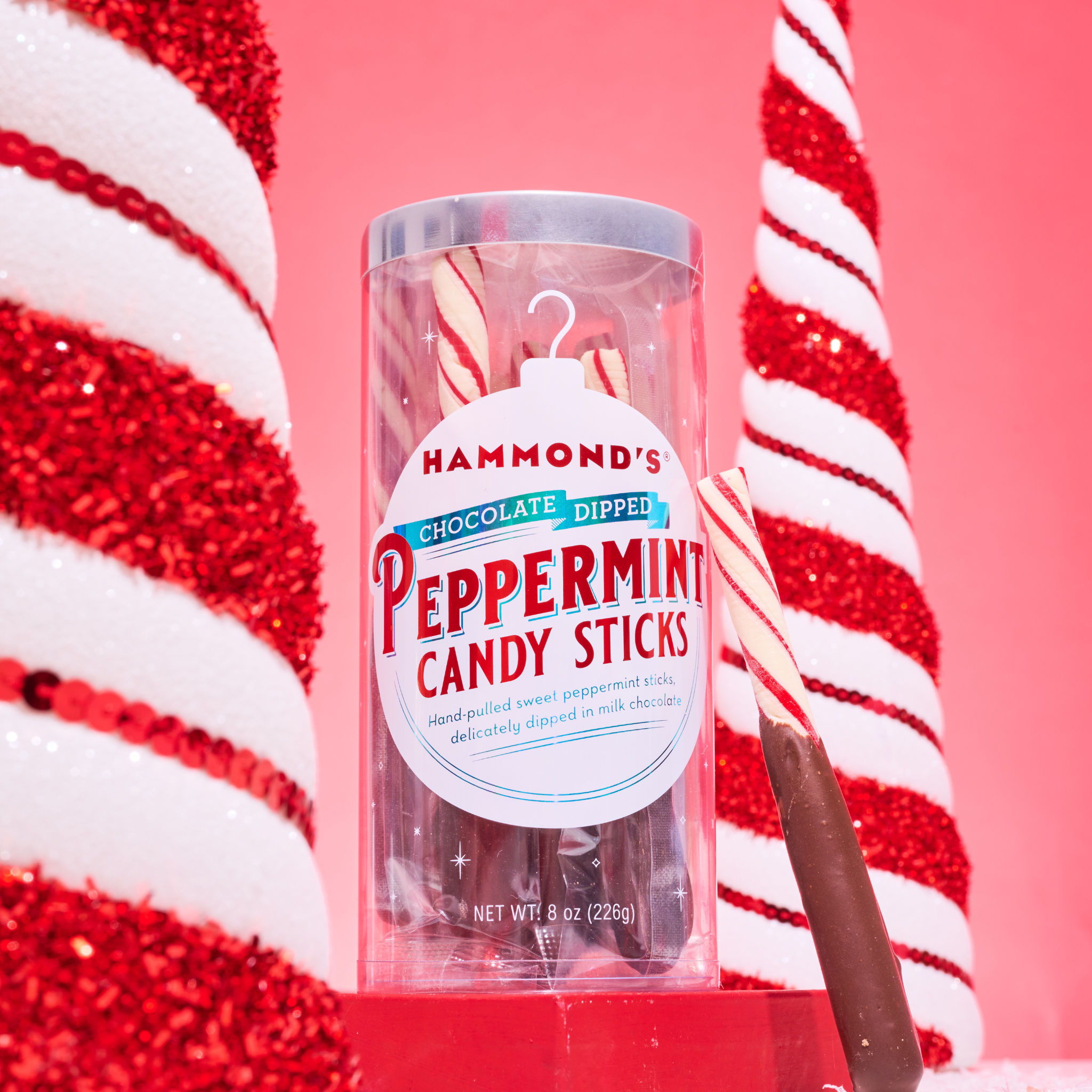 A clear plastic tube packaging containing chocolate-dipped peppermint candy sticks, with red and white striped decorations in the background.